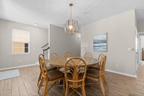 A dining room with a round wooden table, six wicker chairs, a hanging light fixture, and a painting on the wall. - 6-Bedroom Surfside House with Private Pool and Beach Access (Surfside Beach)