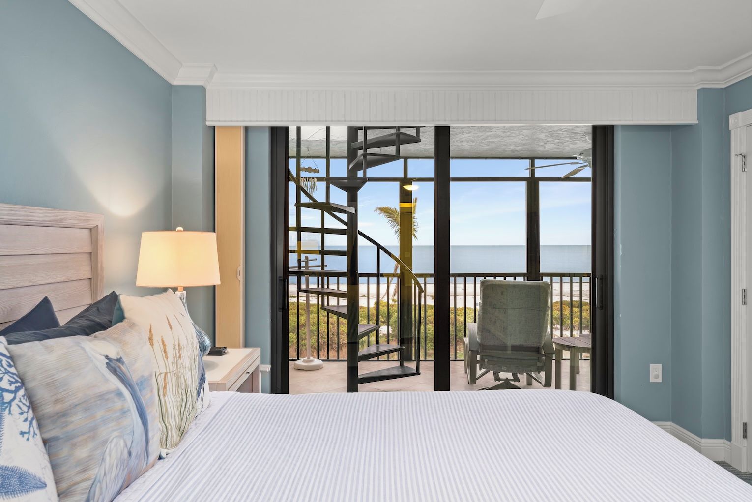 A bedroom with a bed, a nightstand, a lamp, and a view of the beach through a sliding glass door.