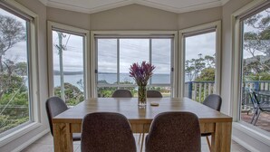 A dining area with a wooden table and four chairs, large windows with a view of the sea, and a vase with flowers on the table. - Front Row Beachfront (Binalong Bay)