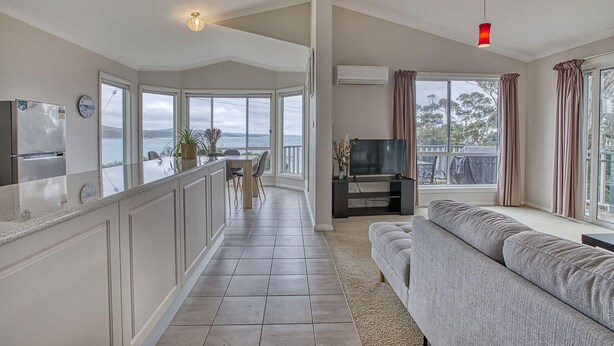 A spacious living area with a kitchen, dining area, and a large window overlooking a body of water. - Front Row Beachfront (Binalong Bay)