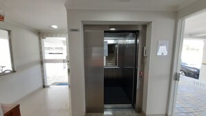Un ascenseur moderne, avec une plaque indiquant « TERREO », se trouve dans un couloir propre et bien éclairé, doté d’un sol carrelé et d’une porte visible donnant sur un espace extérieur.