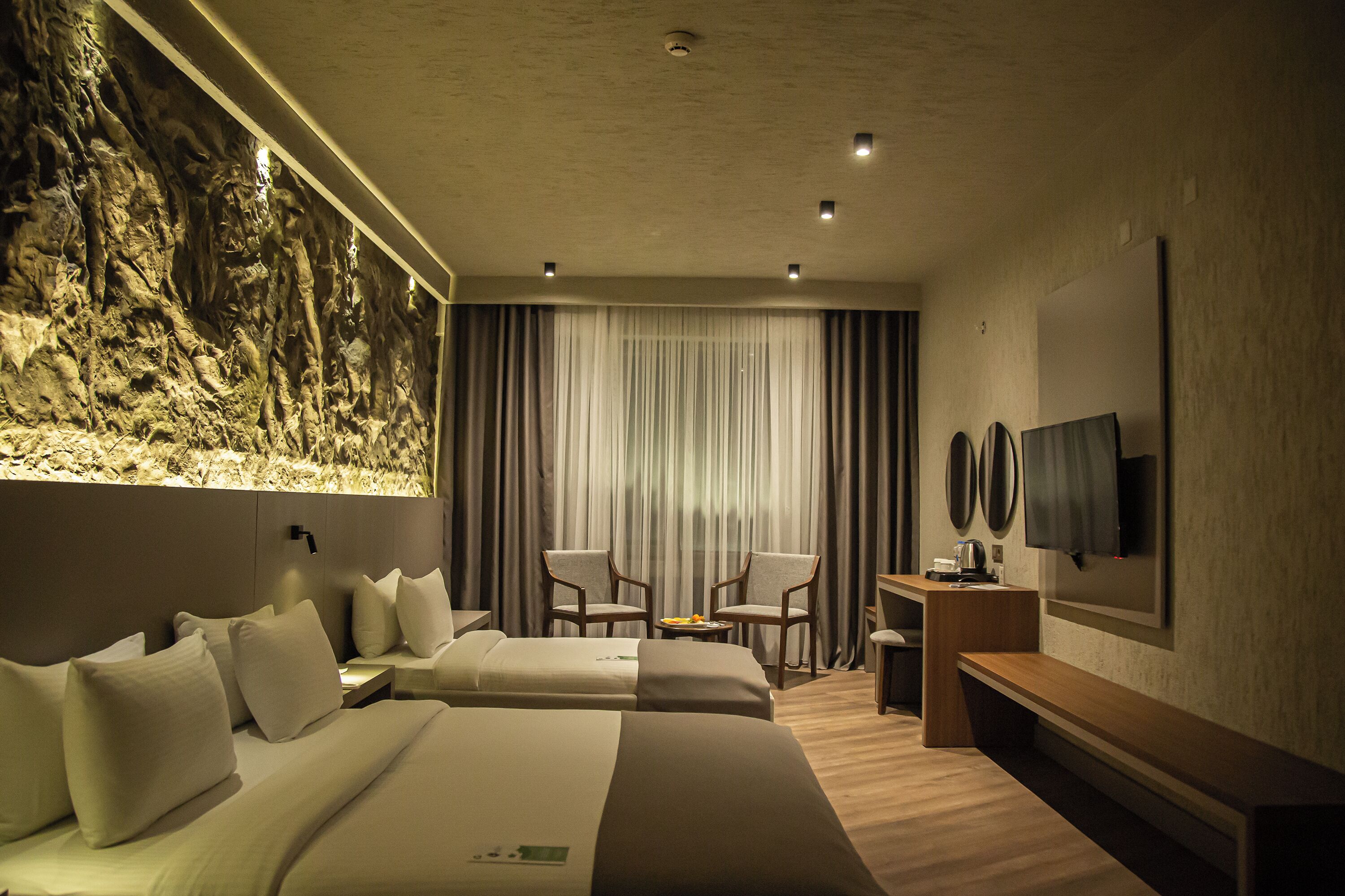 A hotel room with two beds, a desk, chairs, a television, and a large rock wall.