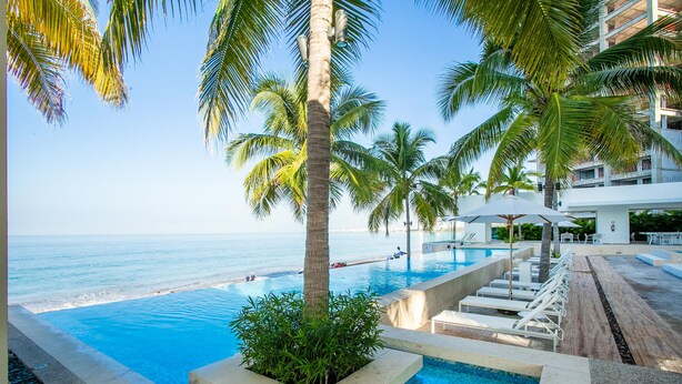 A pool area with clear blue water, palm trees, and a view of the ocean. - Beachfront Luxury Condo, Great Location! (Puerto Vallarta)