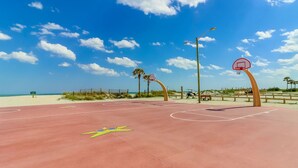Un campo da basket sulla spiaggia con superficie rossa, palme e cielo azzurro limpido.