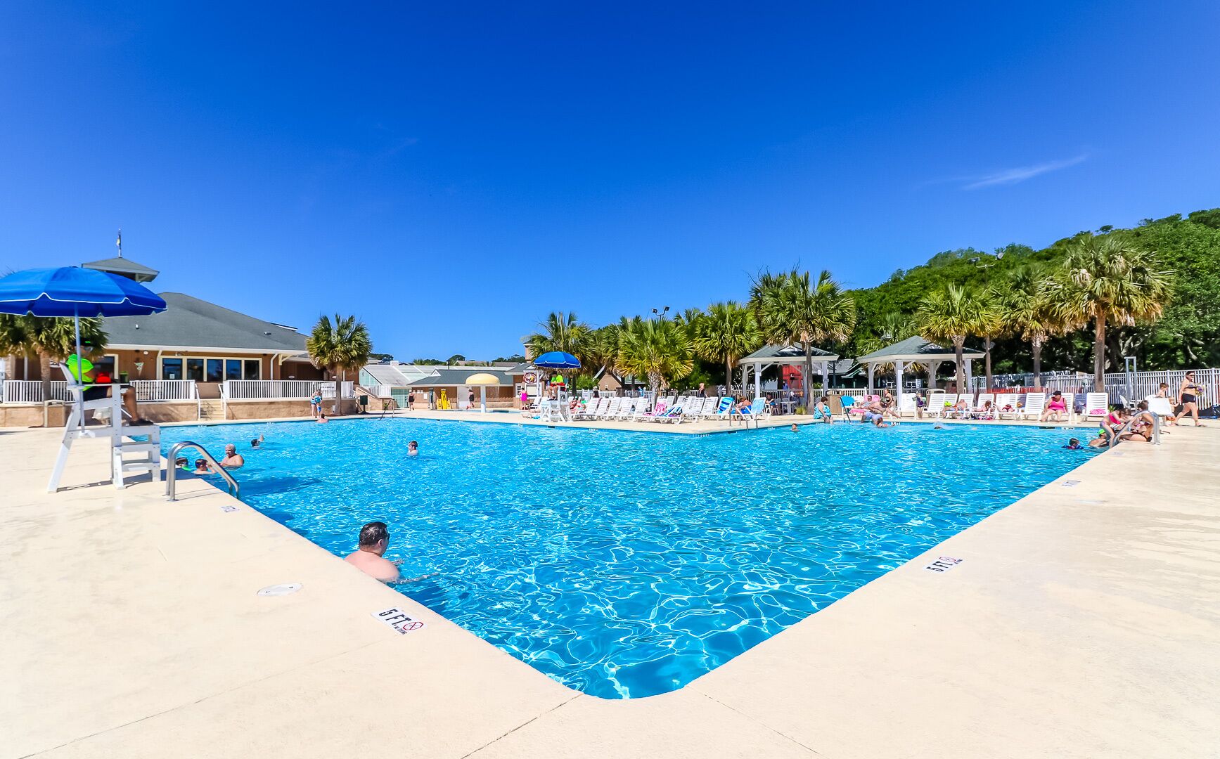 A large outdoor swimming pool with multiple lounge chairs and umbrellas, surrounded by palm trees and a covered pavilion.
