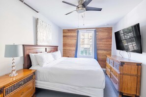 Quarto com cama, cabeceira de madeira, ventilador de teto, televisão, janela com cortinas e cômoda de madeira.