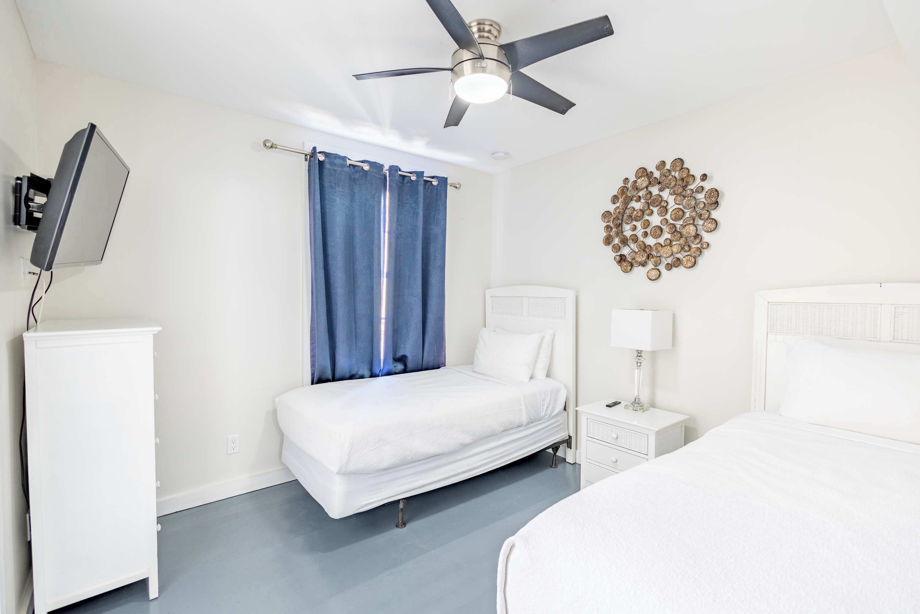A hotel room with a bed, a bedside table with a lamp, a ceiling fan, a TV mounted on the wall, and a decorative wall piece.