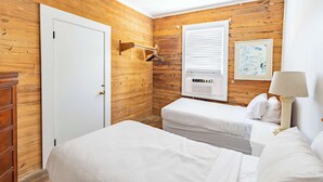 A bedroom with wooden walls, a white door, a window with blinds, a bedside table with a lamp, and a bed. - Commodore's Compound - One block from Duval St (Key West)