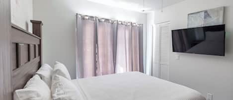 Une chambre à coucher avec un lit, des rideaux, un ventilateur de plafond, un téléviseur fixé au mur et une porte.