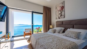 Une chambre avec un grand lit, un bureau et une chaise, un tableau au mur et une vue sur la mer par une porte coulissante.