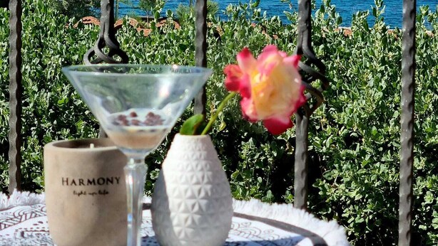 Uma mesa com uma taça de martini, um vaso com uma flor rosa e um castiçal Harmony, com vista para o mar e as montanhas.