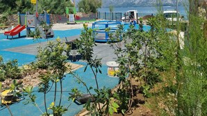 Um parquinho com escorregadores e estruturas de escalada, uma piscina azul e vista para montanhas ao fundo.