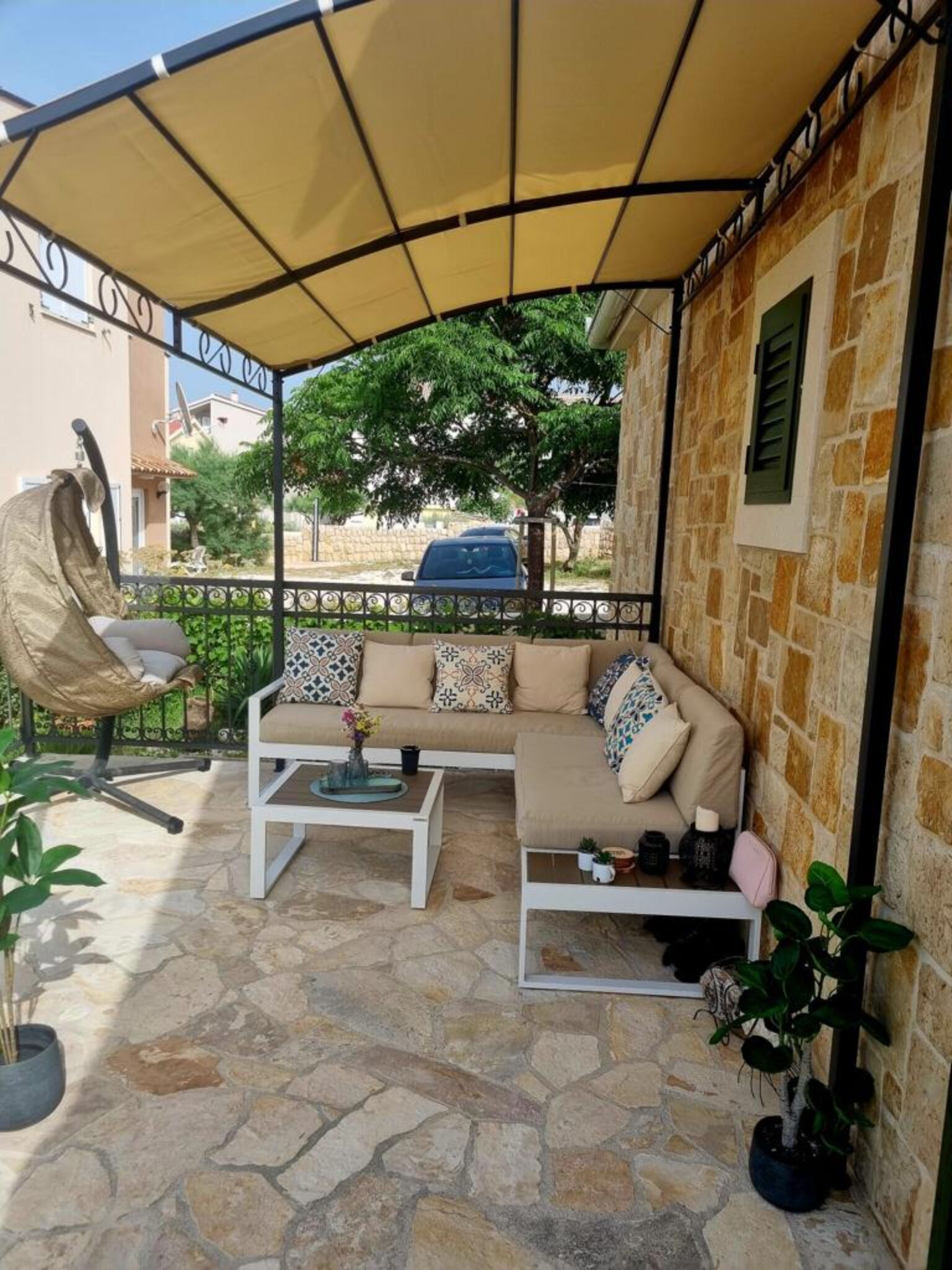 A covered outdoor seating area with a stone floor, a cushioned sofa, a coffee table, and a hanging chair.