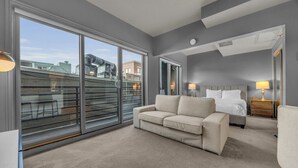 Une chambre d’hôtel moderne dotée d’une grande porte-fenêtre coulissante, d’un canapé beige, d’un lit avec du linge de lit blanc et d’une table de chevet avec une lampe.