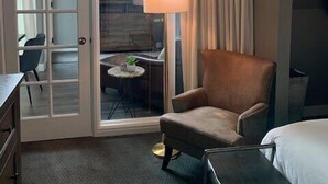 Ein Hotelzimmer mit Bett, Sessel, Schreibtisch und Lampe. Ein großes Fenster mit Vorhängen bietet Blick auf einen anderen Raum.