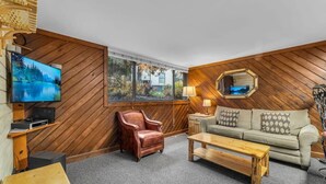 A modern living room with a wooden wall, a sofa, a coffee table, a leather armchair, a TV, and a large window with a view of trees and a house.