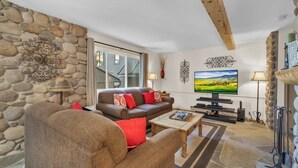 A cozy living room with a stone wall, a brown sofa with red cushions, a wooden coffee table, and a flat-screen TV.