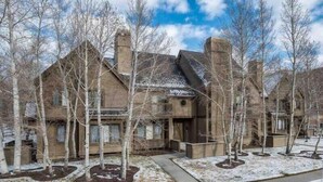 A large, rustic-style house with a prominent chimney, surrounded by a snow-covered landscape and bare trees. - Charming 2 BDR Getaway w/ Views & Private Hot Tub! (Park City)
