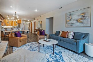 A modern living room with a blue sofa, a coffee table, and a dining area with wooden cabinets and a kitchen in the background. - 3BDR Condo Heart of Old Town Walk to Town Lift (Park City)