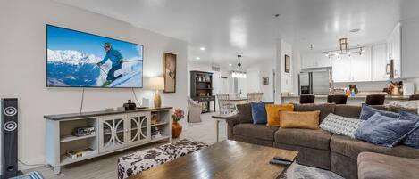 Una sala de estar moderna con un televisor de pantalla plana grande, una mesa de centro de madera y un sofá con varios cojines.