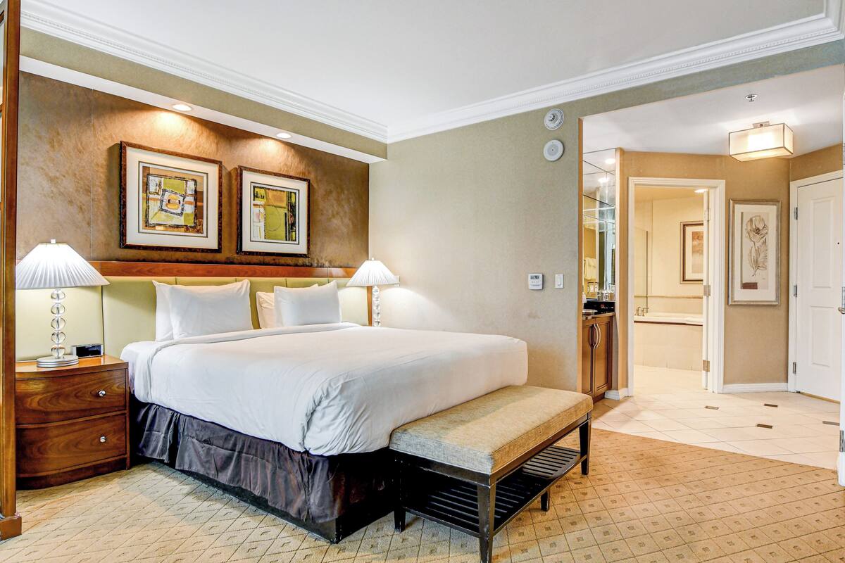 Une chambre d’hôtel avec un grand lit, des tables de chevet avec des lampes, un banc et des œuvres d’art encadrées accrochées au mur.