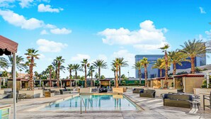 Zona de piscina con sillas de descanso y palmeras, un edificio grande al fondo y un cielo despejado.