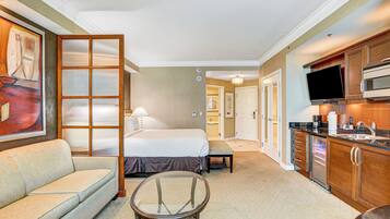 A hotel room with a bed, a sofa, a glass table, a kitchenette, and a painting on the wall.