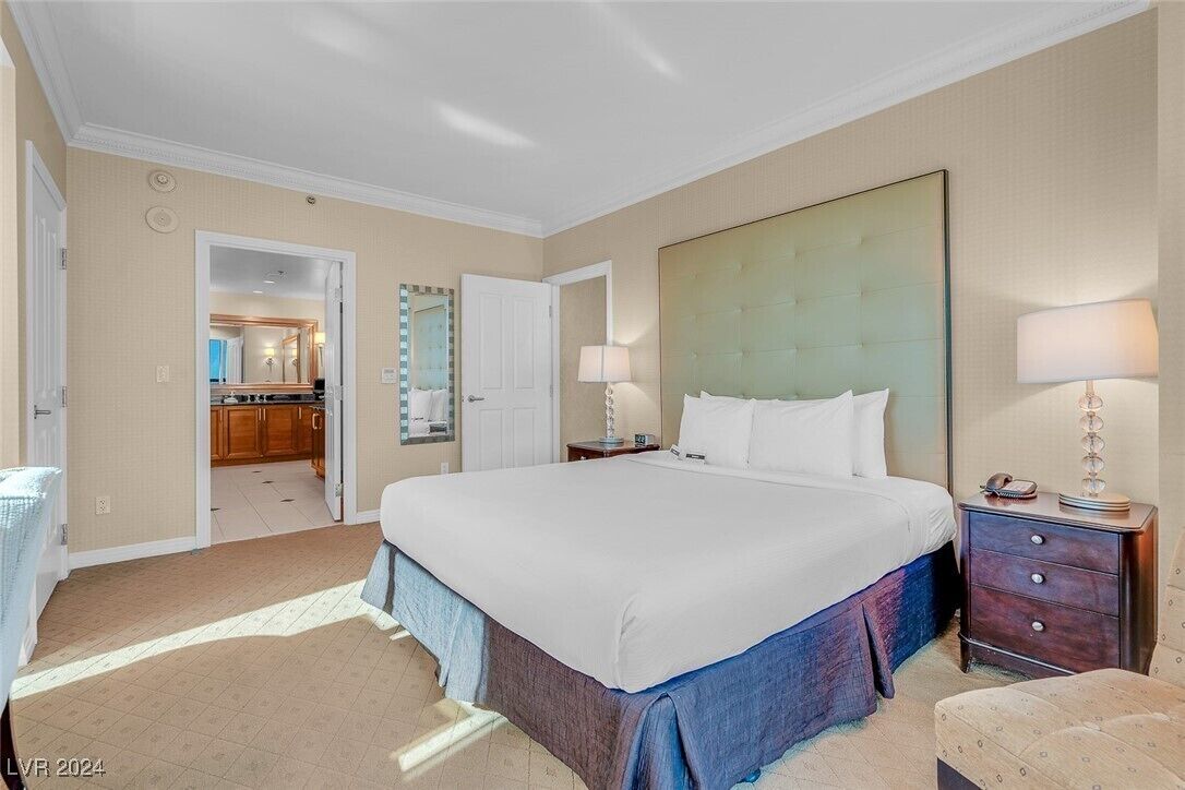 A hotel room with a large bed, a nightstand with a lamp, a dresser, and a view of the bathroom and kitchen through an open door.