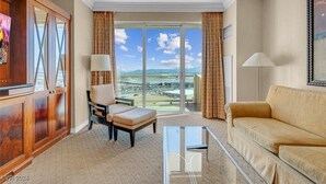 Habitación de hotel con un gran ventanal corredero que da a un balcón con vistas a la playa y las montañas. Hay un sofá, una silla y una mesita con una lámpara.