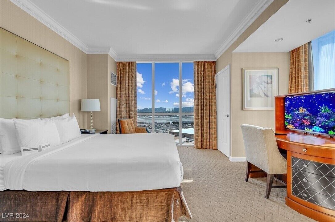 A hotel room with a large bed, a desk with a television, and a view of the city through large windows.