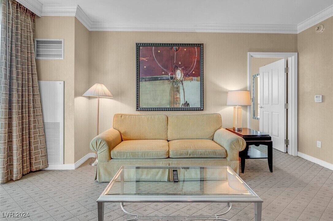 Un salon avec un canapé beige, une table basse en verre, un tableau au mur, une lampe et une porte.