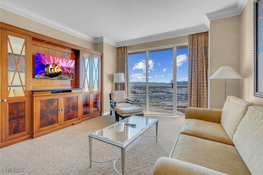 A spacious living room with a large flat-screen TV, a sofa, a glass coffee table, and a view of a cityscape through large windows.