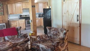 A cozy living room with a fireplace, a ceiling fan, and a kitchen area visible through an open door. - Whitney Landing: A Lakeside Getaway on Upper Cold Stream Pond (Lincoln)