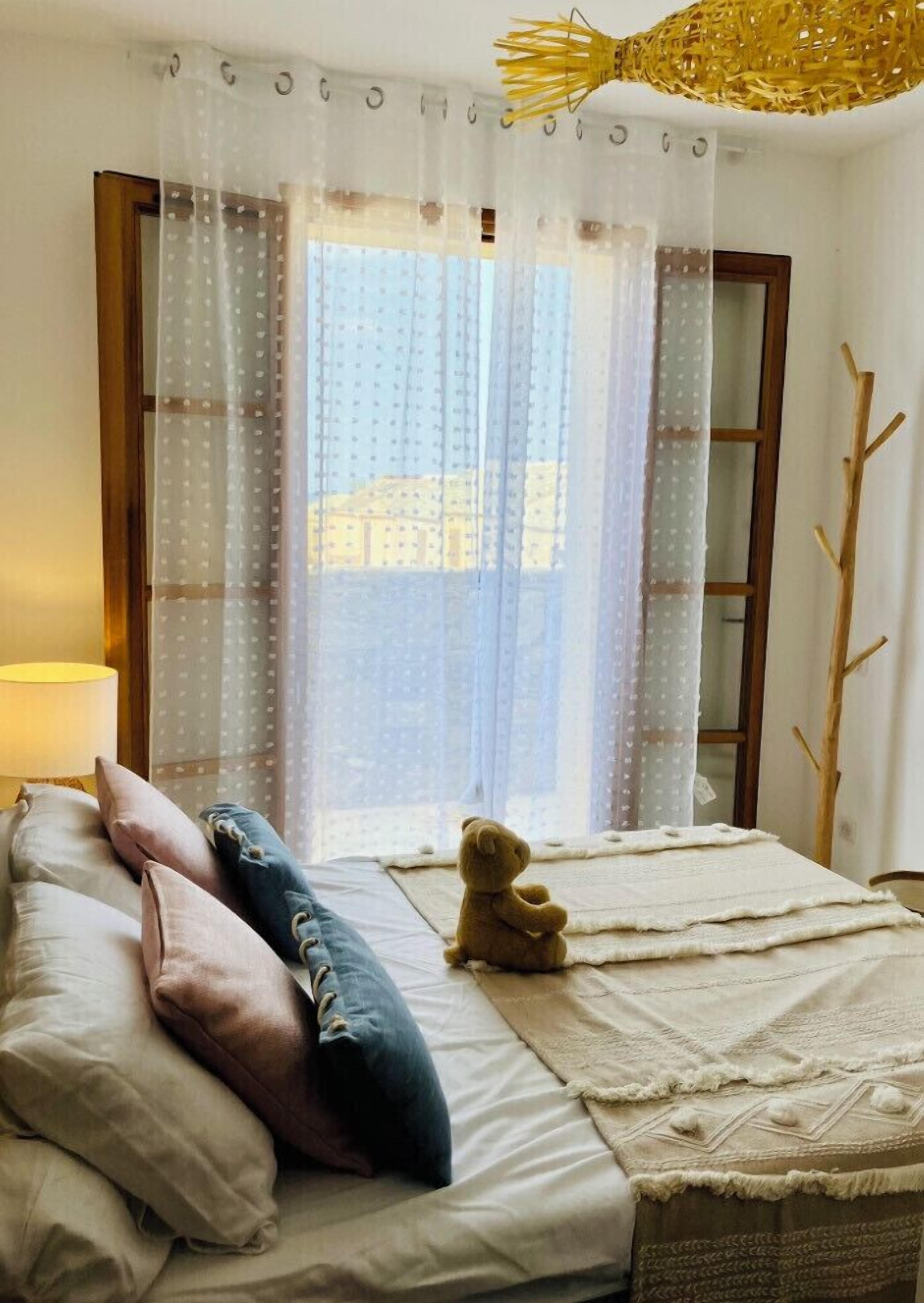 Et soveværelse med en seng, puder, en bamse, en lampe og et vindue med gardiner.