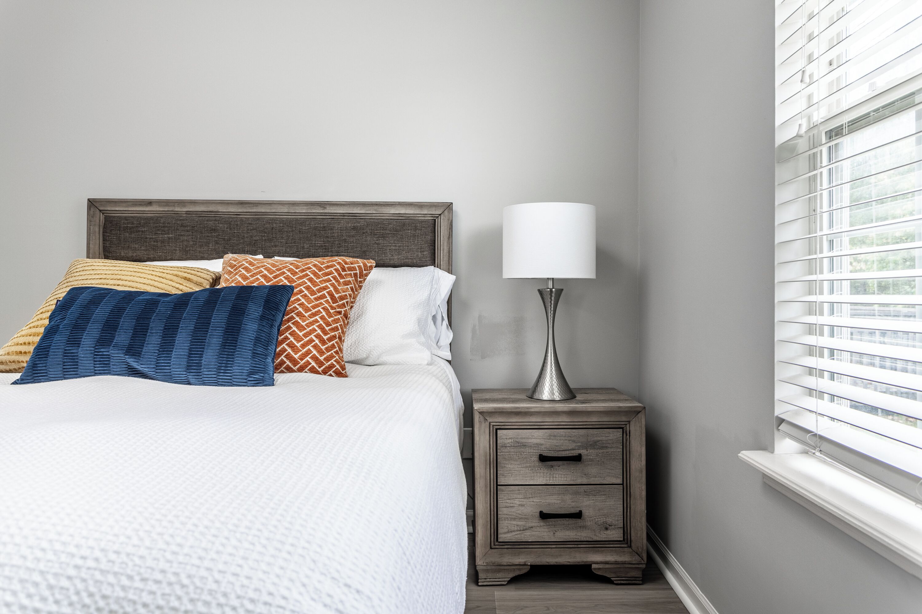 Una cama bien hecha con varios almohadones, una mesita de noche con lámpara y una ventana con persianas.