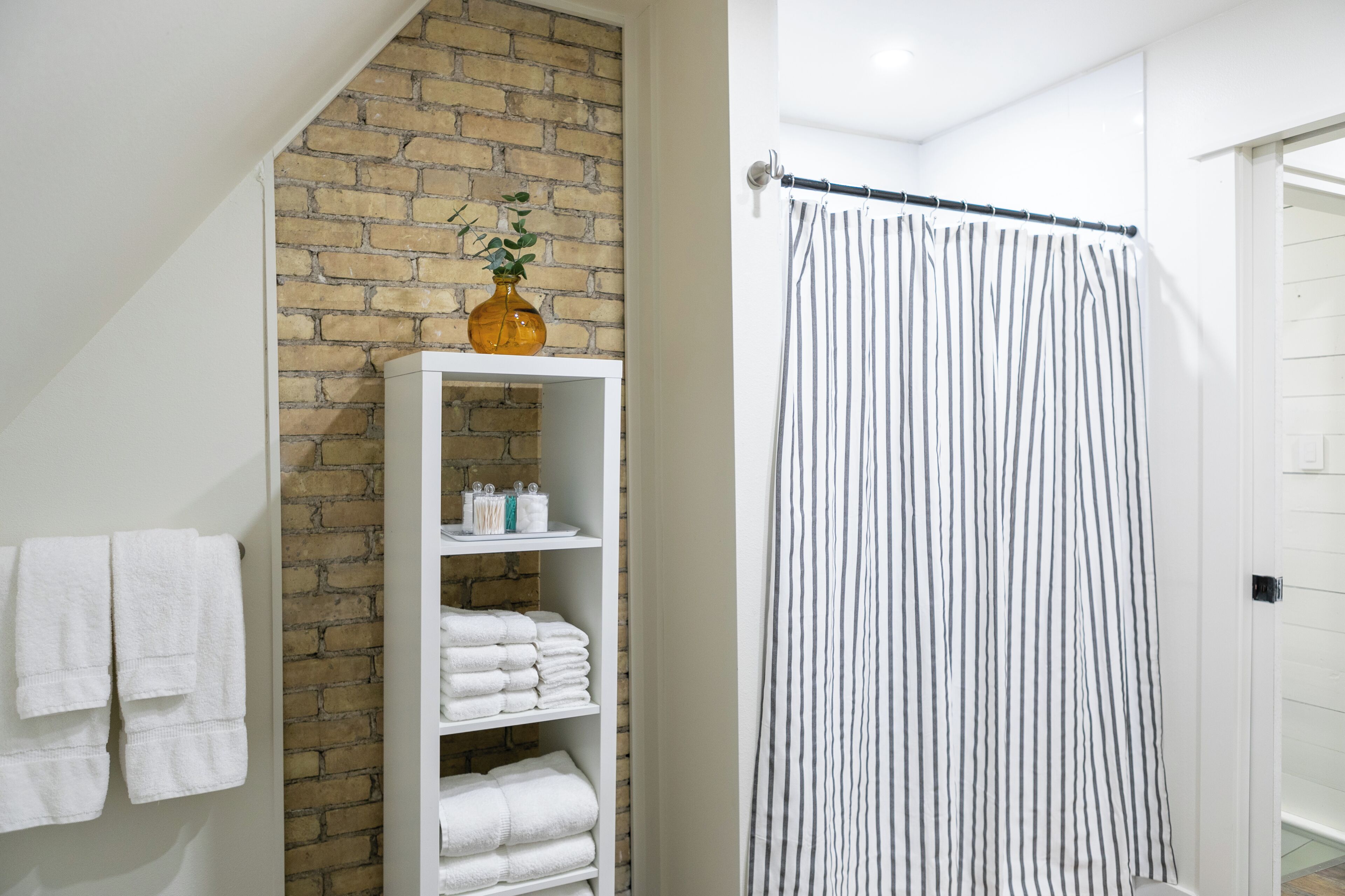 Une salle de bain avec un porte-serviettes blanc, une étagère blanche où sont posées des serviettes, un rideau de douche à rayures et un mur en briques.