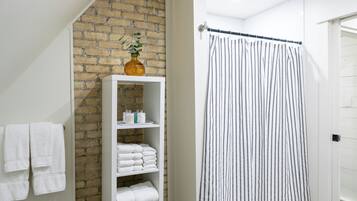 Une salle de bain avec un porte-serviettes blanc, une étagère blanche où sont posées des serviettes, un rideau de douche à rayures et un mur en briques.