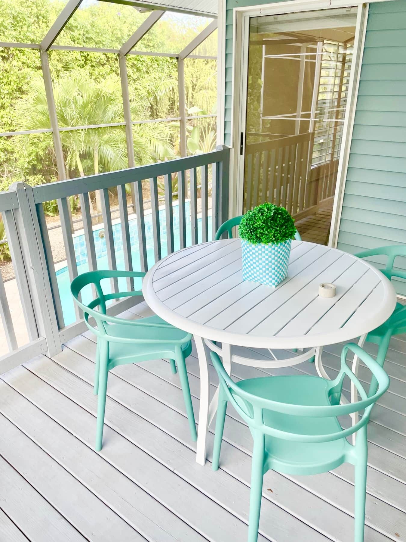 A covered outdoor area with a round table and four chairs, a potted plant, and a view of a swimming pool.