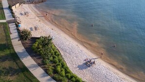 Vista aérea de uma praia com calçadão pavimentado, um molhe rochoso e pessoas aproveitando a água.
