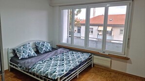 Un letto singolo con copriletto e cuscini decorati, un termosifone sotto la finestra e vista sugli edifici.