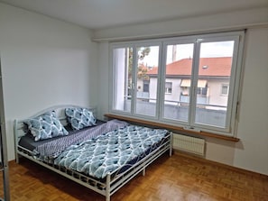 Uma cama de solteiro com colcha e almofadas estampadas, um radiador abaixo da janela e vista para prédios.