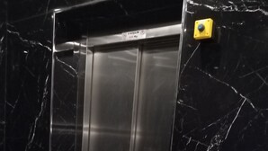 Un ascensor modern amb acabat metàl·lic, un panell de control amb un botó groc i una paret de marbre amb un dibuix de fissures.
