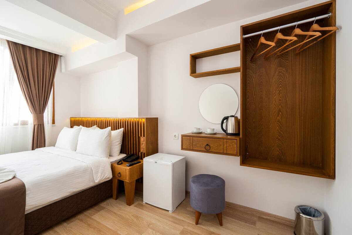 A modern hotel room with a bed, a small refrigerator, a wooden wardrobe, a mirror, and a bedside table with a lamp.