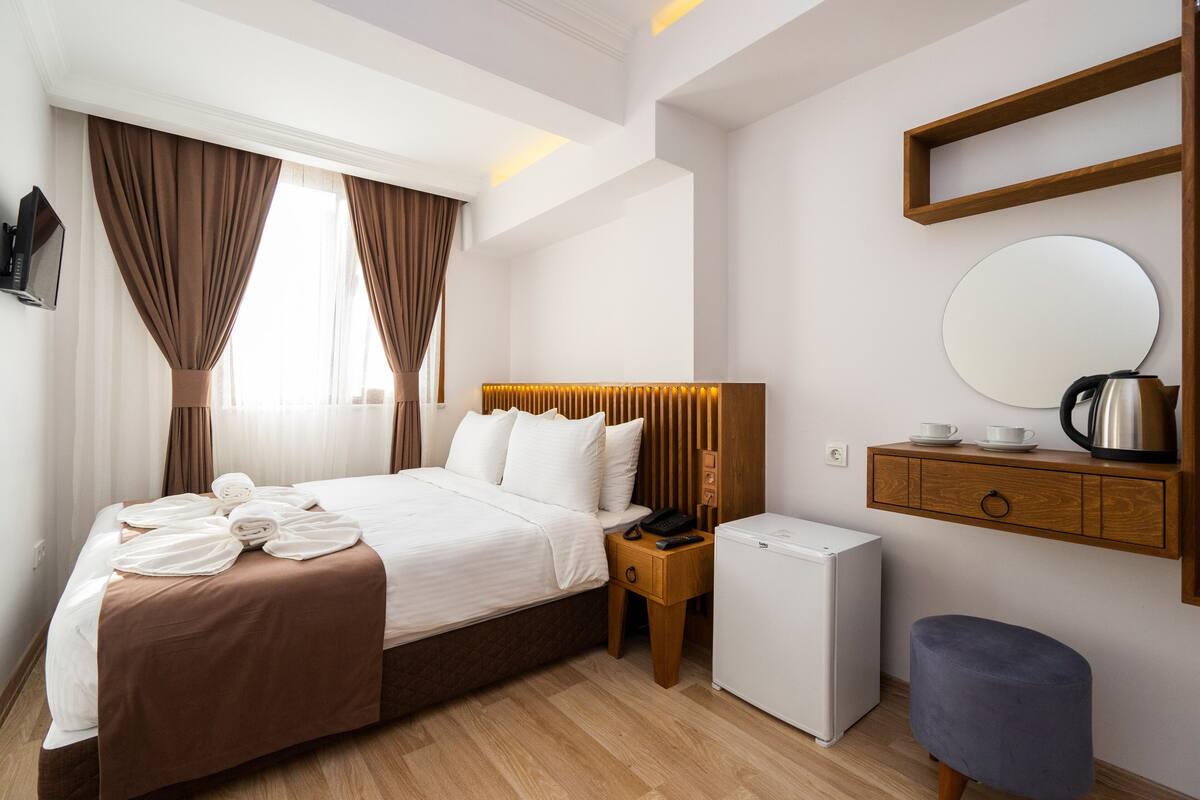 A hotel room with a bed, a small refrigerator, a wooden nightstand, a blue stool, a mirror, and a shelf with a kettle and cups.