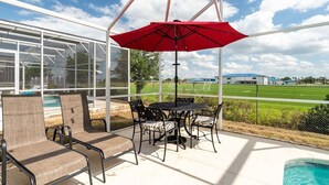 Eine Terrasse im Freien mit Tisch und Stühlen, einem roten Sonnenschirm und Blick auf ein Schwimmbecken und grüne Felder.