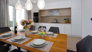 A modern dining area with a wooden table set for four, white plates, cutlery, and a vase with flowers. A kitchen is visible in the background with white cabinets and a fruit bowl. - Villa Marko (Makarska)