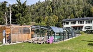 A glass-enclosed pool area with lounge chairs, a wooden structure, and a building with solar panels in the background. - Chalet Capucine 1 in the heart of the Vosges mountains (Gérardmer)
