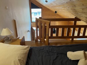 Ein Schlafzimmer mit Holzbalkendecke, einem Bett mit dunkler Bettdecke, einem Nachttisch mit Lampe und Blick durch ein Fenster.