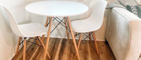 Una habitación pequeña y acogedora con una mesa redonda blanca y dos sillas blancas, un sofá color crema, y una ventana con persianas.