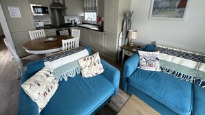 A cozy living room with a blue sofa set, a wooden dining table, and a kitchen area with white cabinets and a stainless steel range hood. - Stunning Coastal Beach House - Sleeping 5 in 2 Bedrooms - Pool and Beach Access Included. (Filey)